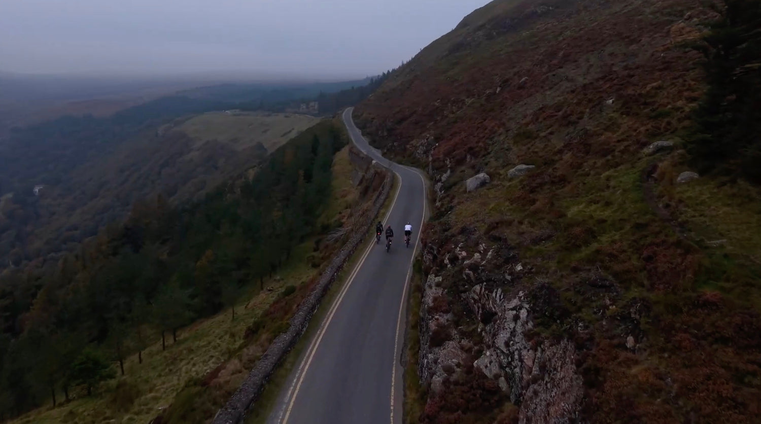 Drone view of cyclists riding along the winding mountain road near Lough Tay, Wicklow, with heather-covered hills stretching into the distance. 